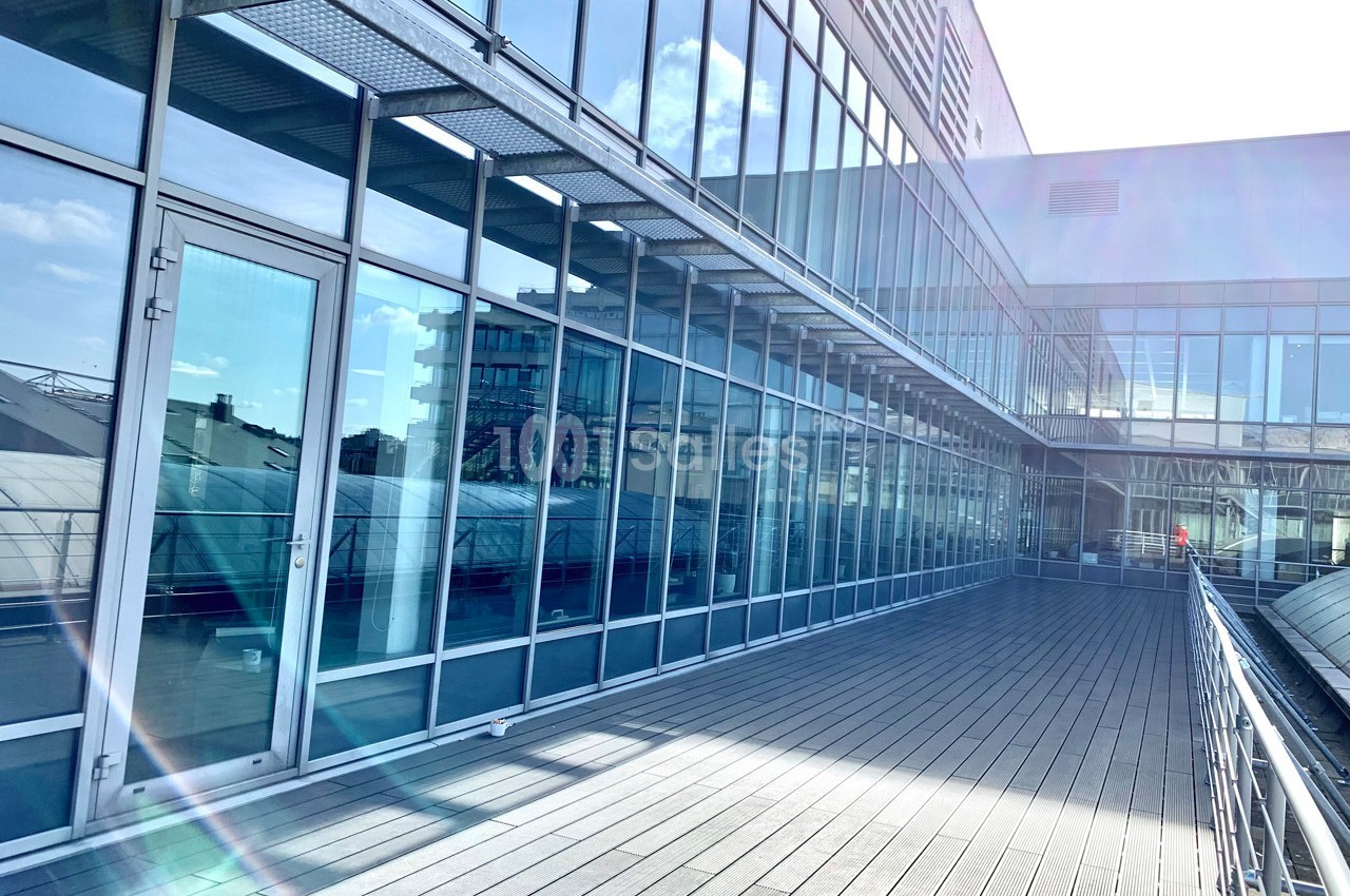 Façade vitrée d'un bâtiment moderne avec une terrasse en bois éclairée par le soleil.