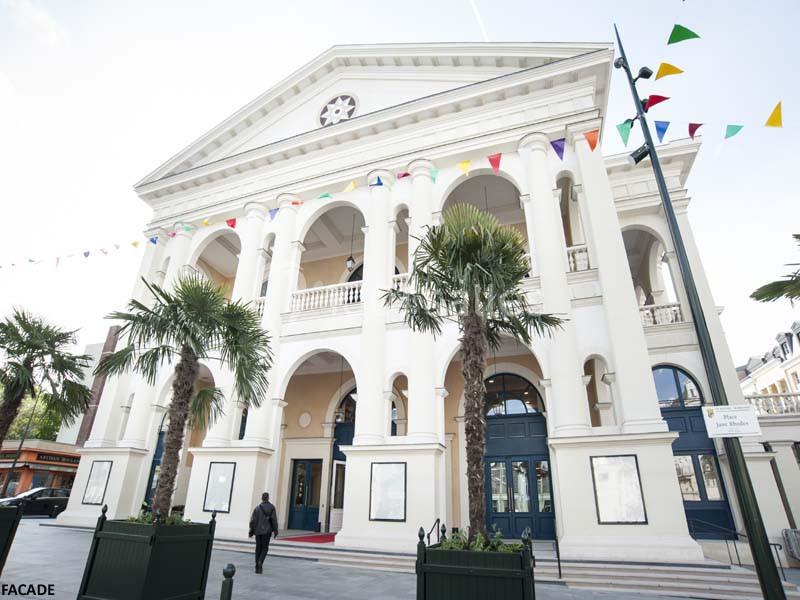Façade d'un bâtiment néoclassique avec colonnes, décorée de fanions colorés, entourée de palmiers et d'un passant.