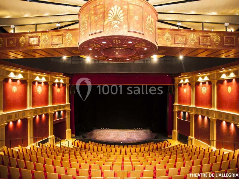 Salle de théâtre avec des rangées de sièges rouges, une scène sombre et un plafond orné de détails dorés.