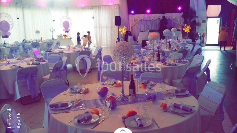 Salle de réception décorée pour un événement, avec tables rondes dressées, nappes blanches et éclairage coloré.