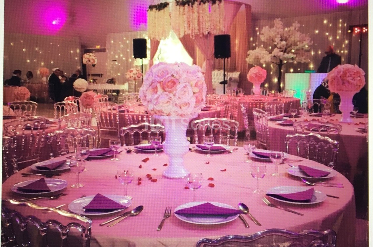 Salle décorée pour un événement avec tables rondes dressées, nappes roses, centres de table fleuris et éclairage tamisé.