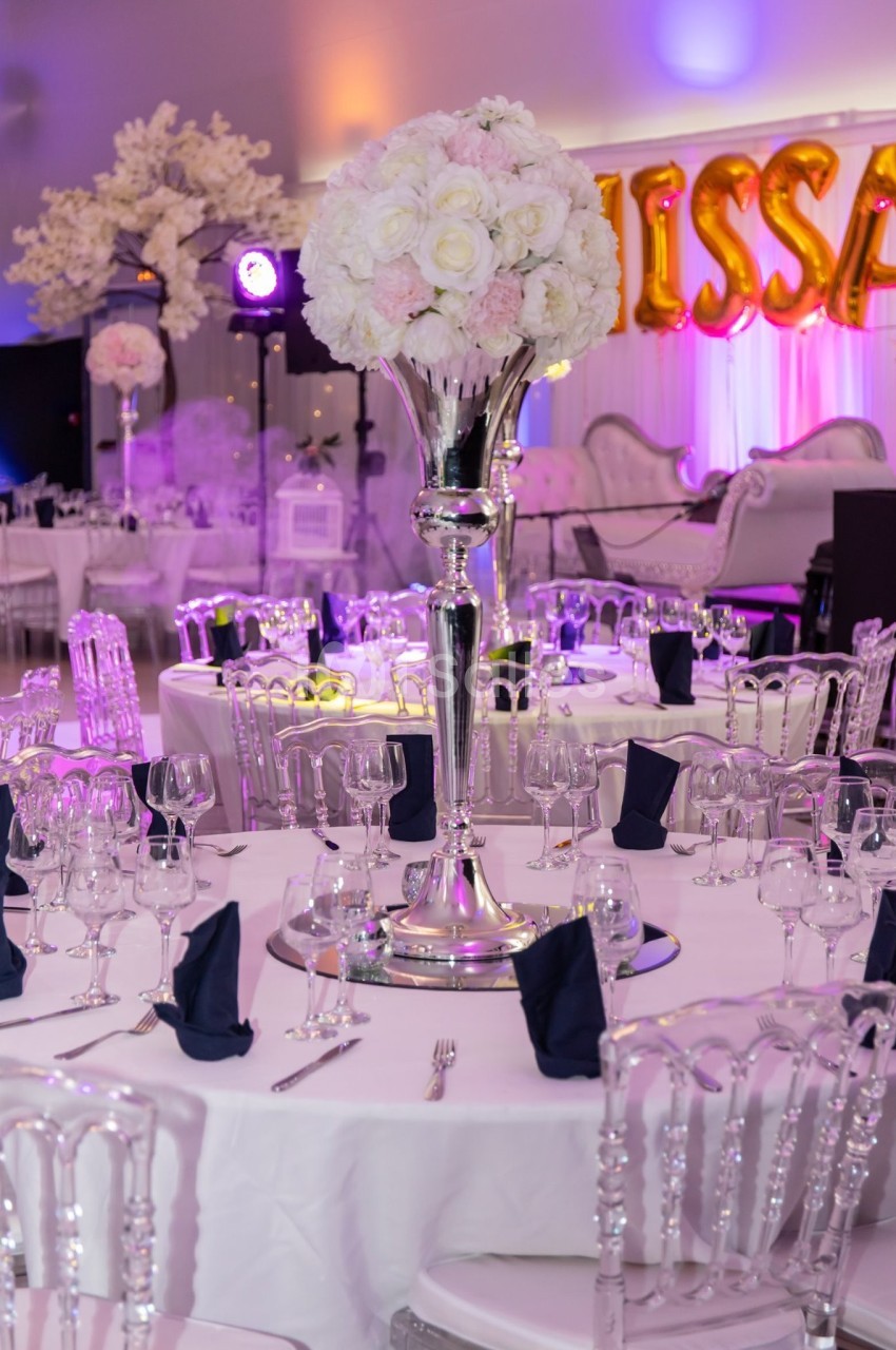 Salle de réception décorée avec des tables élégantes, des chaises transparentes et un centre de table floral sur pied…