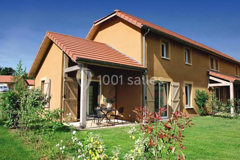 Maison avec façade ocre, toit en tuiles rouges, terrasse aménagée et jardin verdoyant sous un ciel bleu.