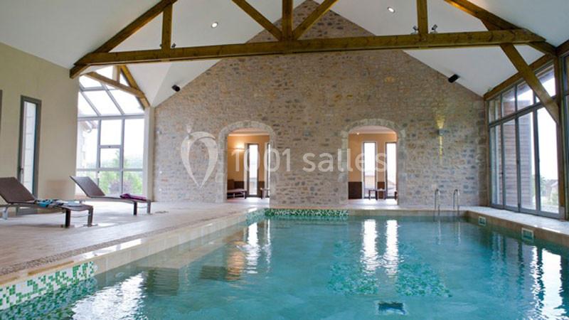 Piscine intérieure dans une pièce en pierre avec poutres apparentes, entourée de chaises longues et grandes fenêtres.
