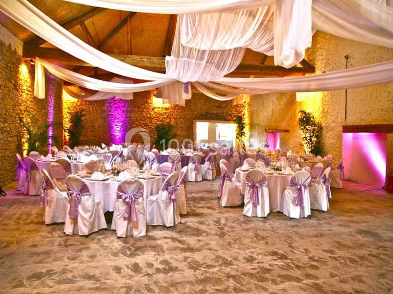 Salle de réception décorée avec des tables rondes, nappes blanches, chaises ornées de nœuds violets et voilages suspendus.