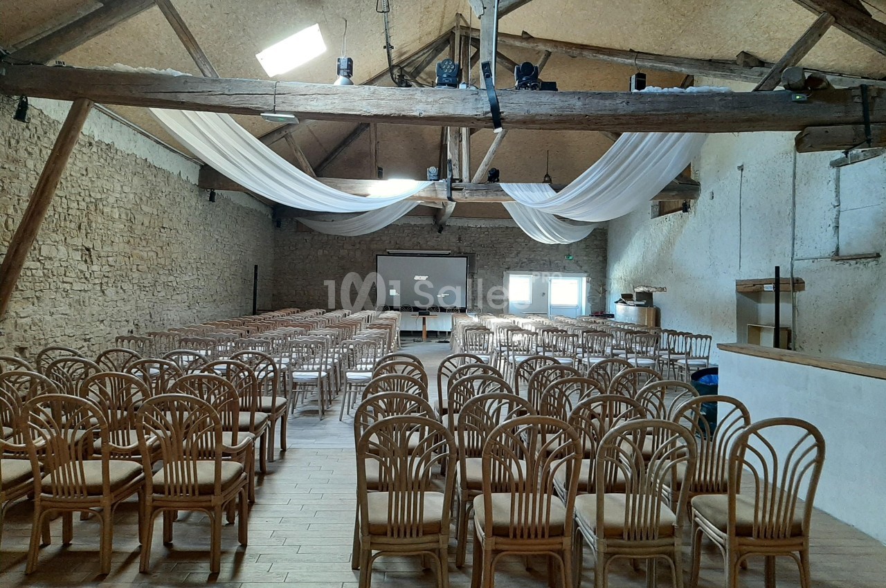 Salle de réception avec des rangées de chaises en bois, un écran de projection et des poutres apparentes au plafond.