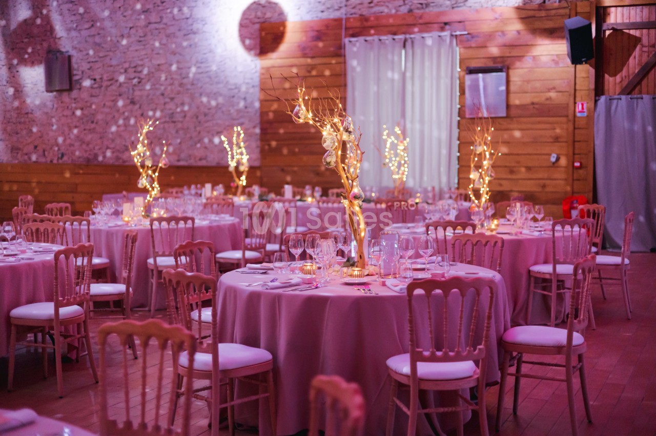 Salle de réception décorée avec des tables rondes, nappes roses, chaises en bois et centres de table lumineux.