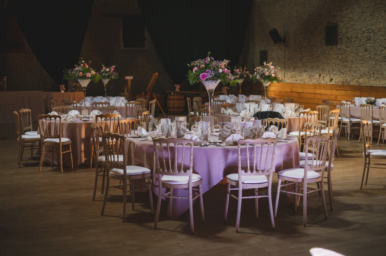 Salle de réception décorée avec des tables rondes nappées de rose, chaises en bois clair et arrangements floraux.