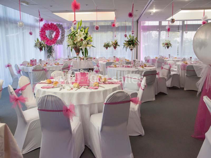 Salle décorée pour un événement, avec des tables rondes dressées, des housses blanches et des accents roses.