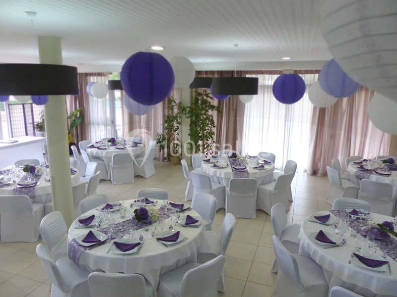 Salle de réception décorée avec des tables rondes, nappes blanches, serviettes violettes et lanternes suspendues.