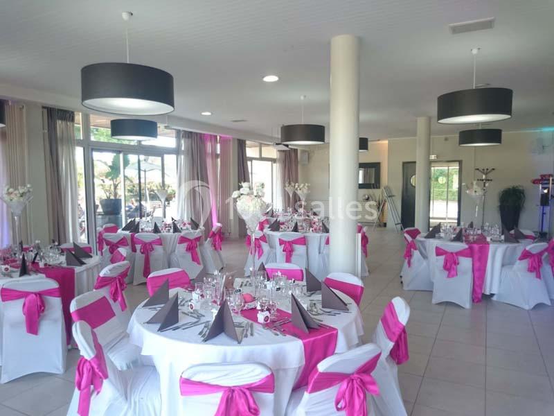 Salle de réception lumineuse avec tables rondes décorées de nappes blanches et nœuds roses sur les chaises.