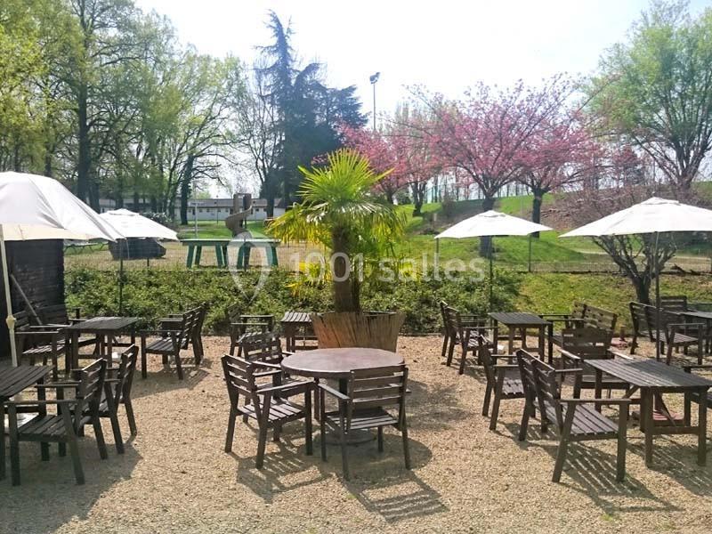 Terrasse extérieure avec tables et chaises en bois, entourée d'arbres et de parasols, par une journée ensoleillée.
