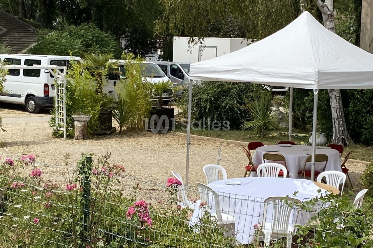 Tables et chaises blanches sous un barnum dans un jardin avec des fleurs, des arbustes et des véhicules en arrière-plan.