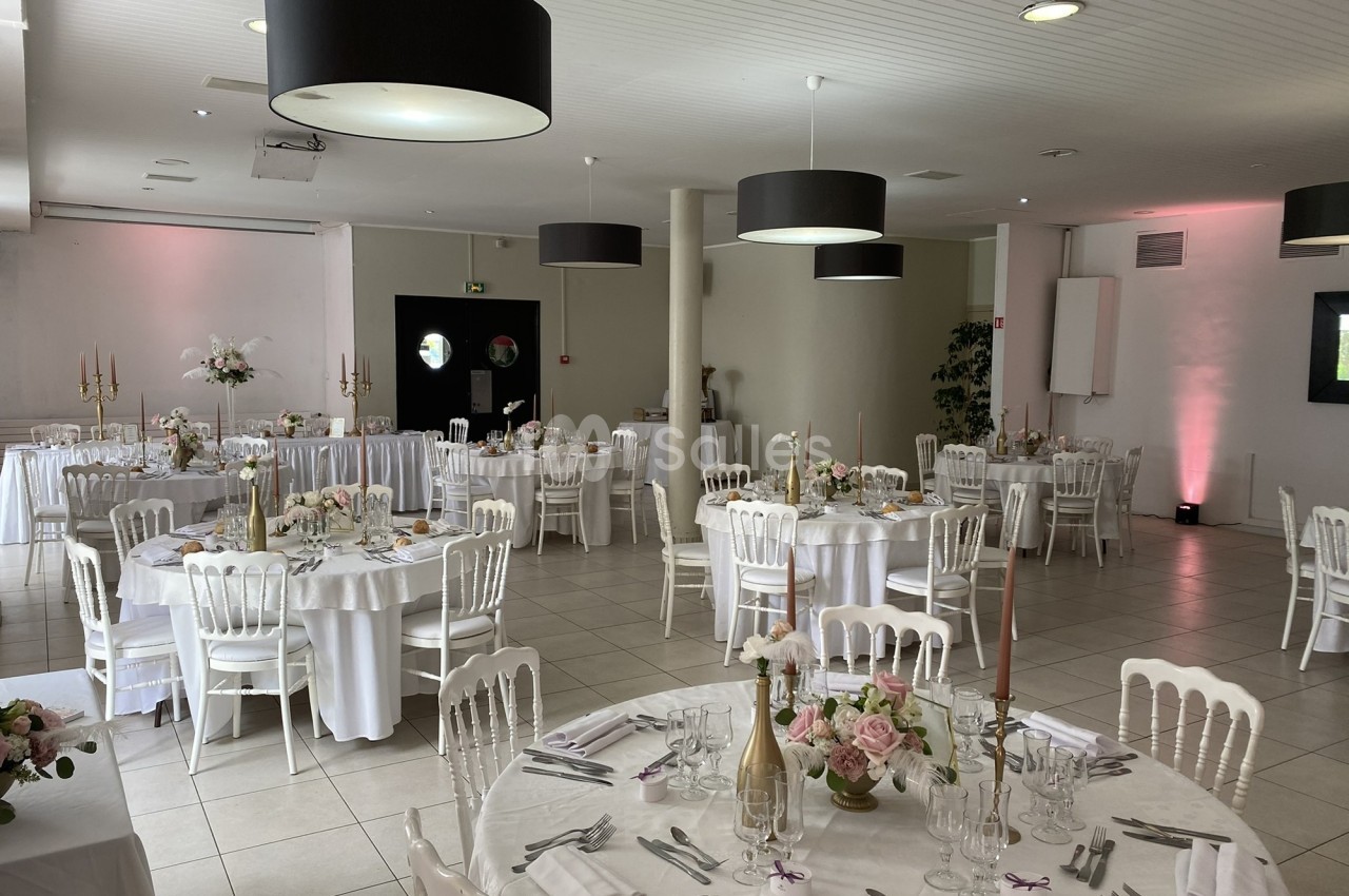 Salle de réception décorée avec des tables rondes, nappes blanches, chaises élégantes et arrangements floraux.