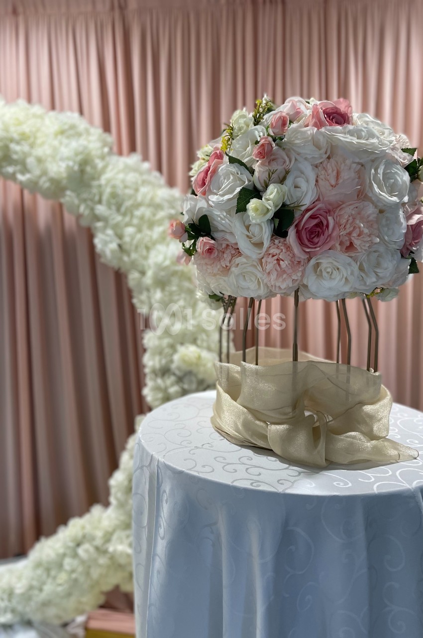 Arrangement floral rond avec des roses blanches et roses sur un support doré, devant un rideau rose et une arche fleurie…