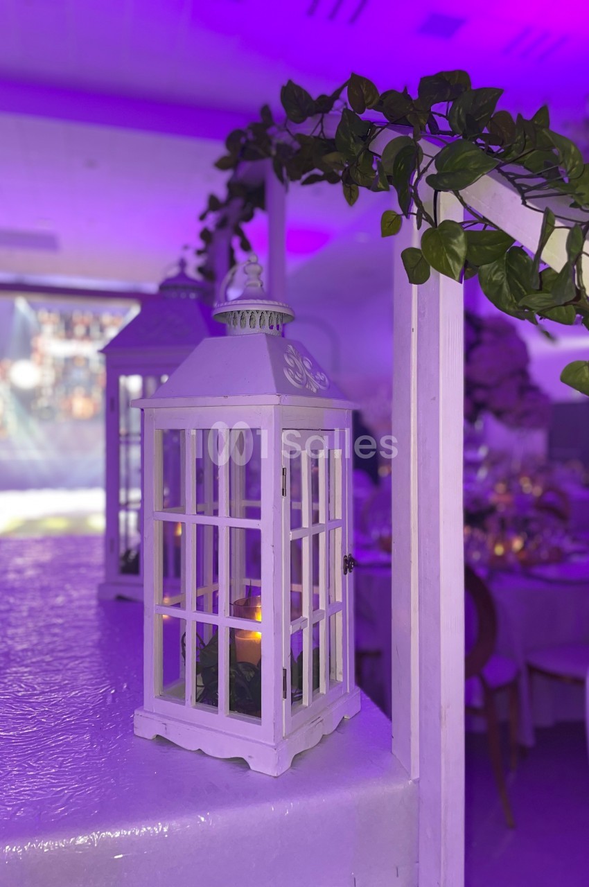 Lanterne blanche décorative avec une bougie allumée, entourée de feuillage, dans une salle éclairée par des lumières…