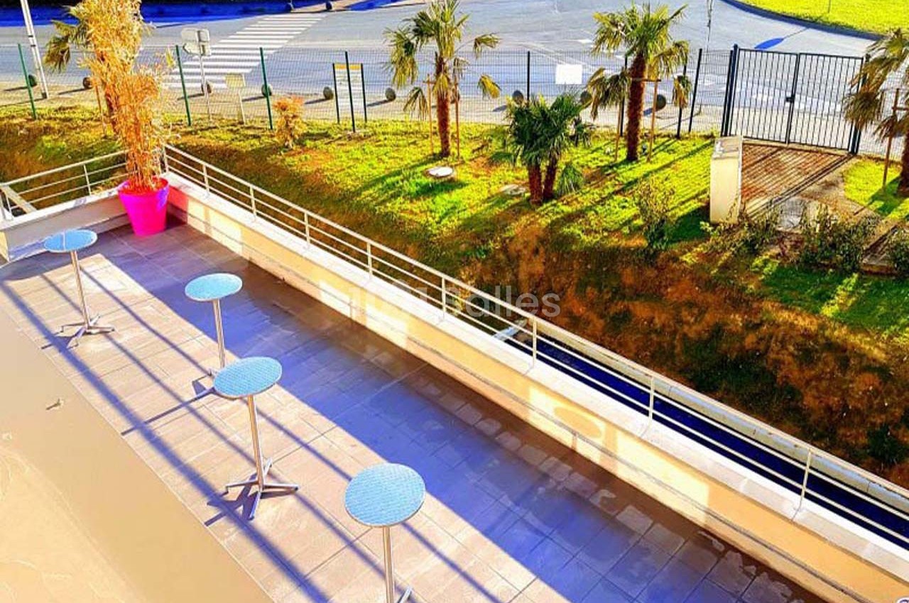 Terrasse ensoleillée avec tables hautes rondes, vue sur un espace vert clôturé et une route en arrière-plan.