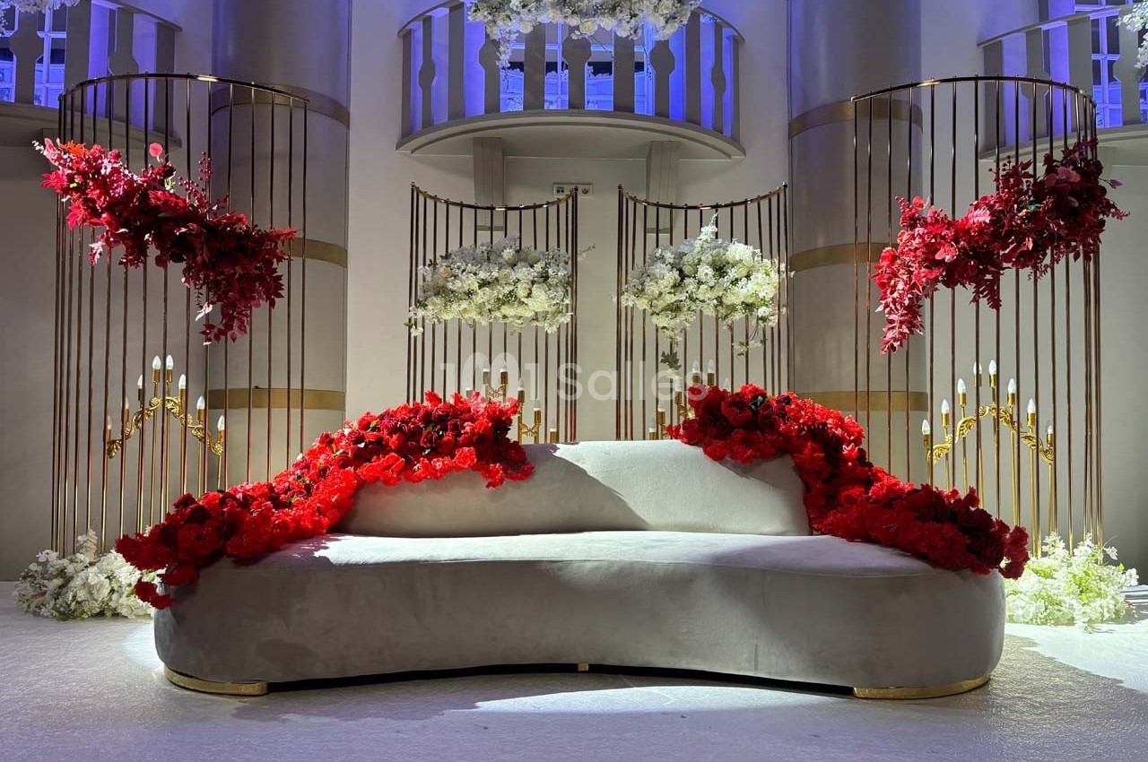 Canapé blanc décoré de fleurs rouges et blanches, entouré de structures dorées ornées de fleurs suspendues.
