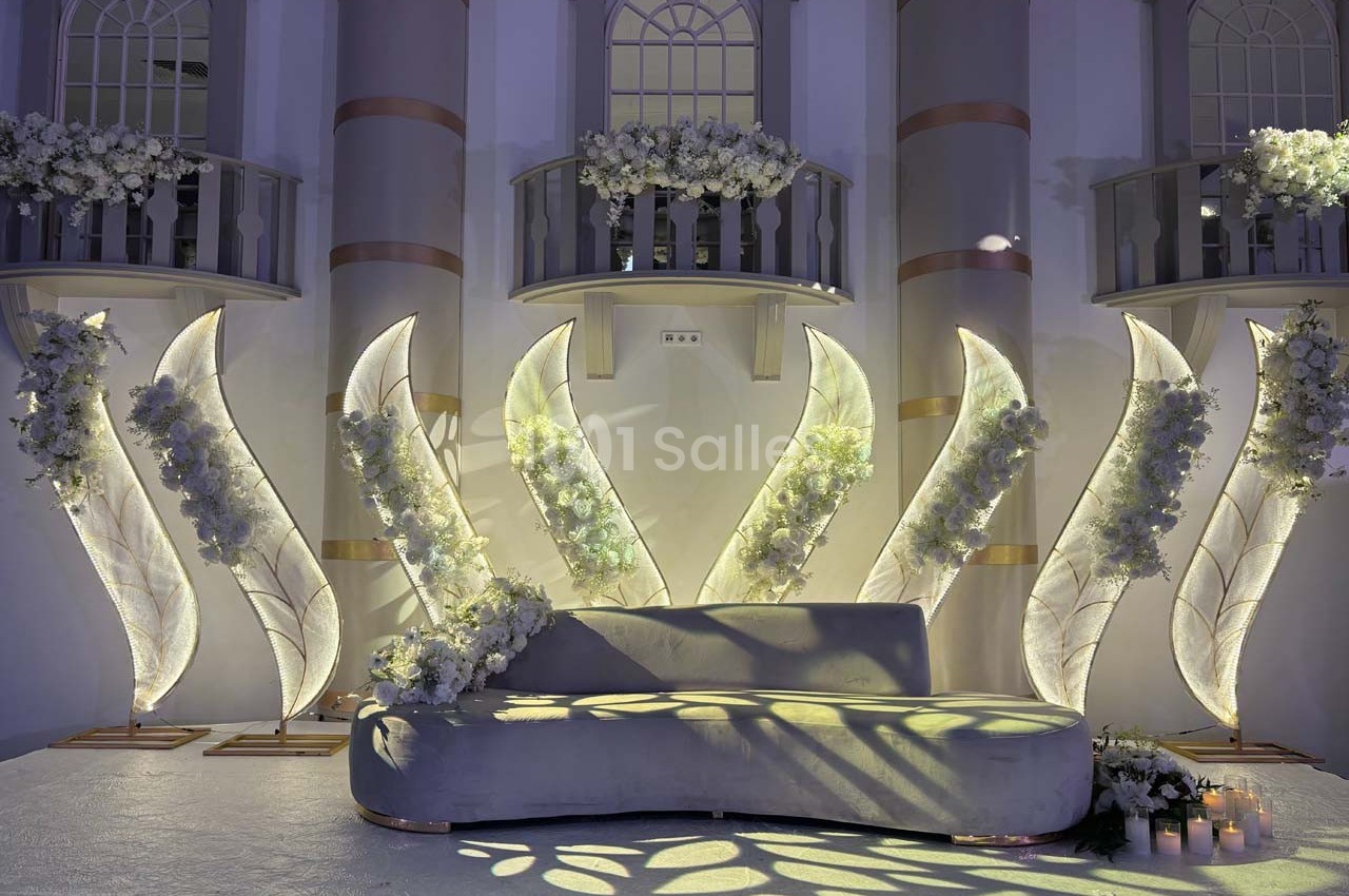 Décor élégant avec un canapé gris, des fleurs blanches et des structures lumineuses en forme de feuilles devant une façade.