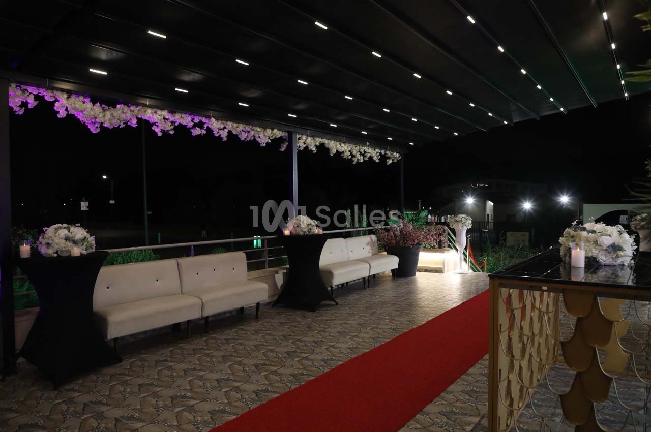 Terrasse éclairée de nuit avec canapé blanc, tables hautes décorées de fleurs et tapis rouge au sol.