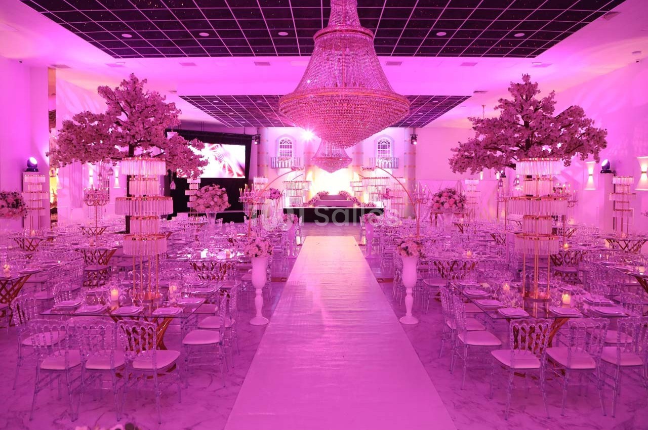 Salle de réception décorée avec des tons roses, des tables élégantes, des chandeliers et un grand lustre central.