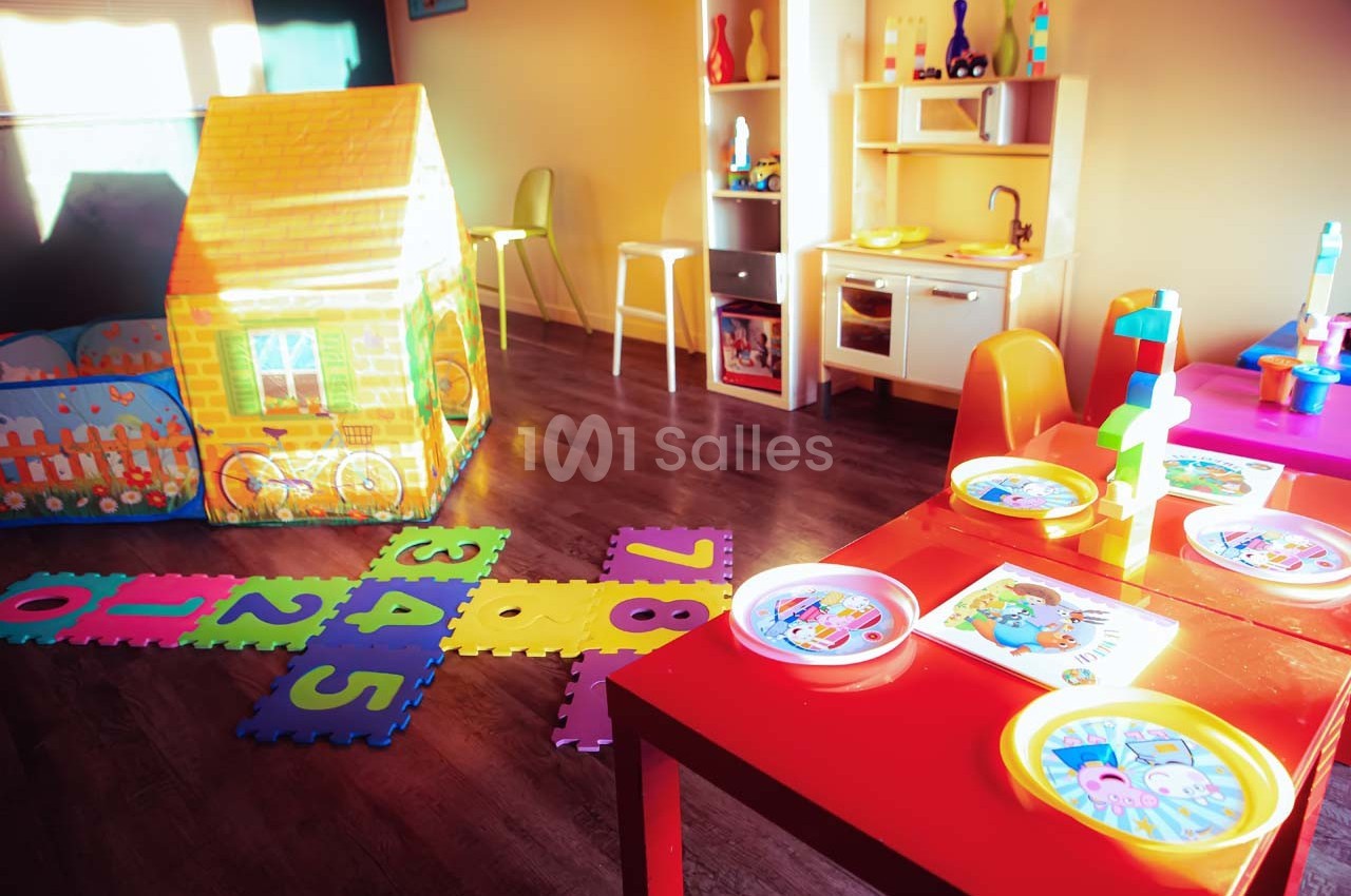 Salle de jeux lumineuse avec une cabane en tissu, un tapis de chiffres, une cuisine pour enfants et des tables colorées.