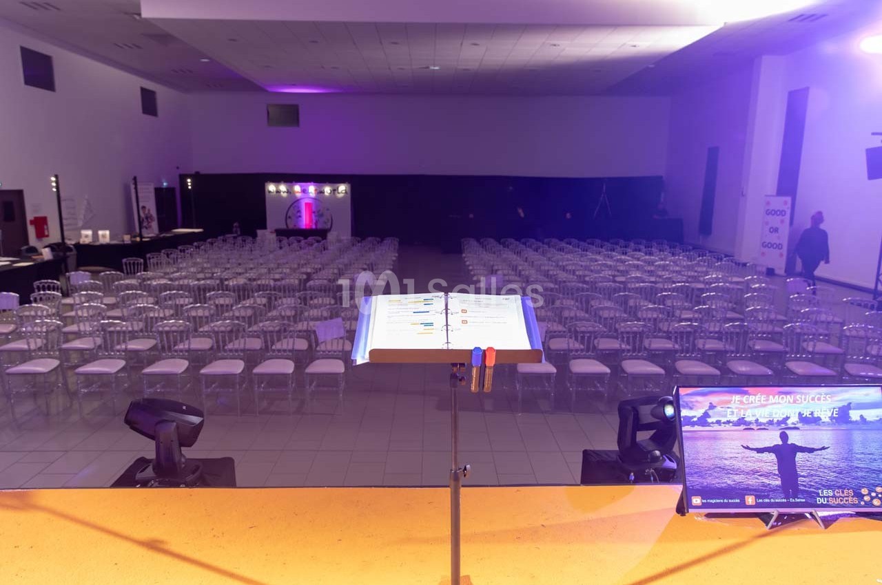 Salle de conférence vide avec des rangées de chaises alignées, vue depuis un pupitre équipé d'un écran et de documents.