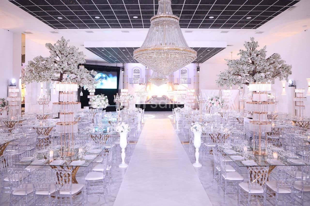 Salle de réception luxueuse avec tables en verre, chaises transparentes, arbres décoratifs et grand lustre central.