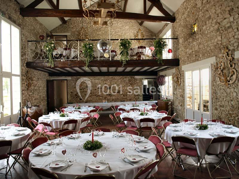 Salle de réception rustique avec murs en pierre, tables rondes dressées et mezzanine décorée de plantes.