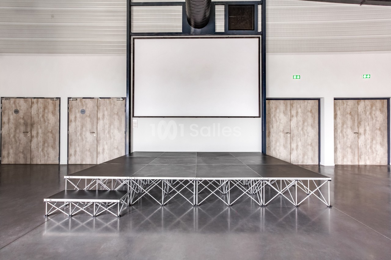 Estrade métallique avec une petite marche, placée dans une salle moderne avec un écran blanc et des portes en bois.