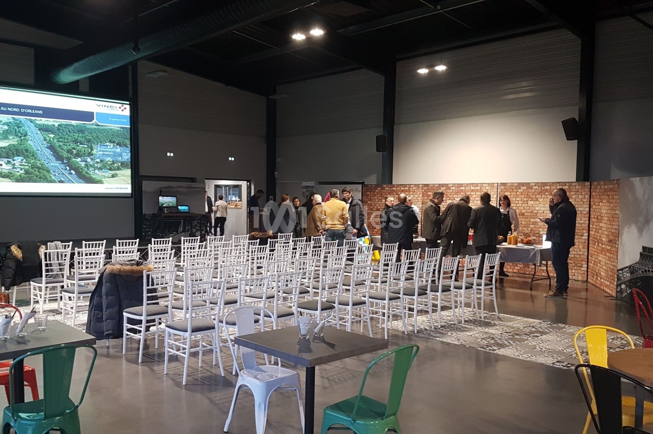 Salle de conférence avec des chaises blanches alignées, un écran de projection et un groupe de personnes discutant au fond.