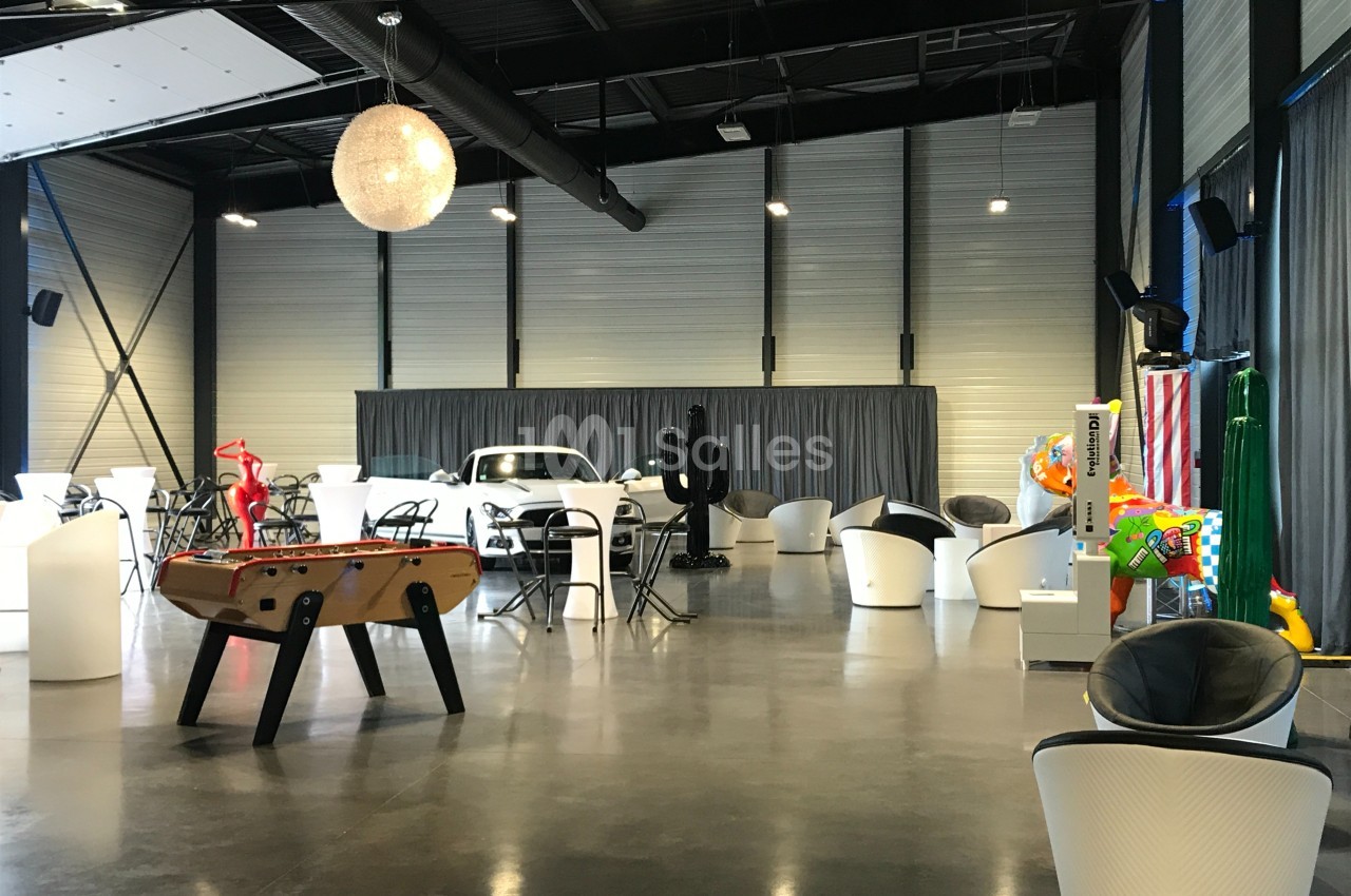 Salle moderne avec sol en béton, mobilier varié, baby-foot, décorations colorées et une voiture blanche en arrière-plan.