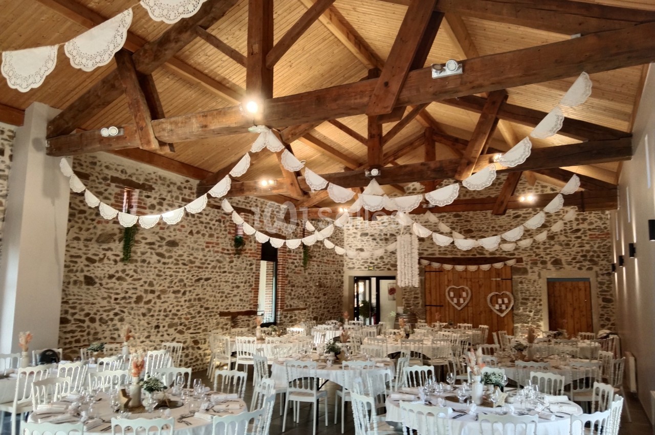 Salle de réception décorée avec des guirlandes blanches, des tables dressées et des chaises blanches sous un plafond en bois.