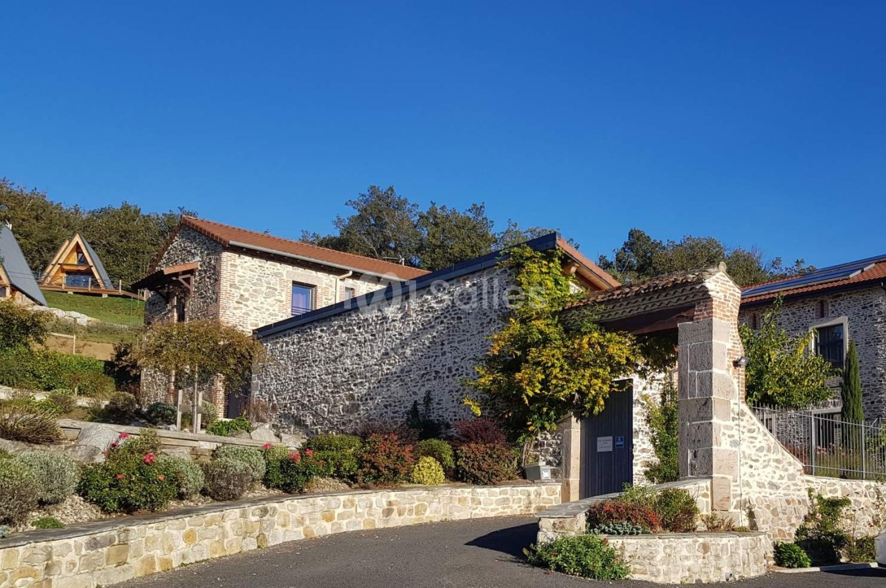 Maison en pierre avec toit en tuiles, entourée de végétation et située dans un environnement rural sous un ciel bleu.