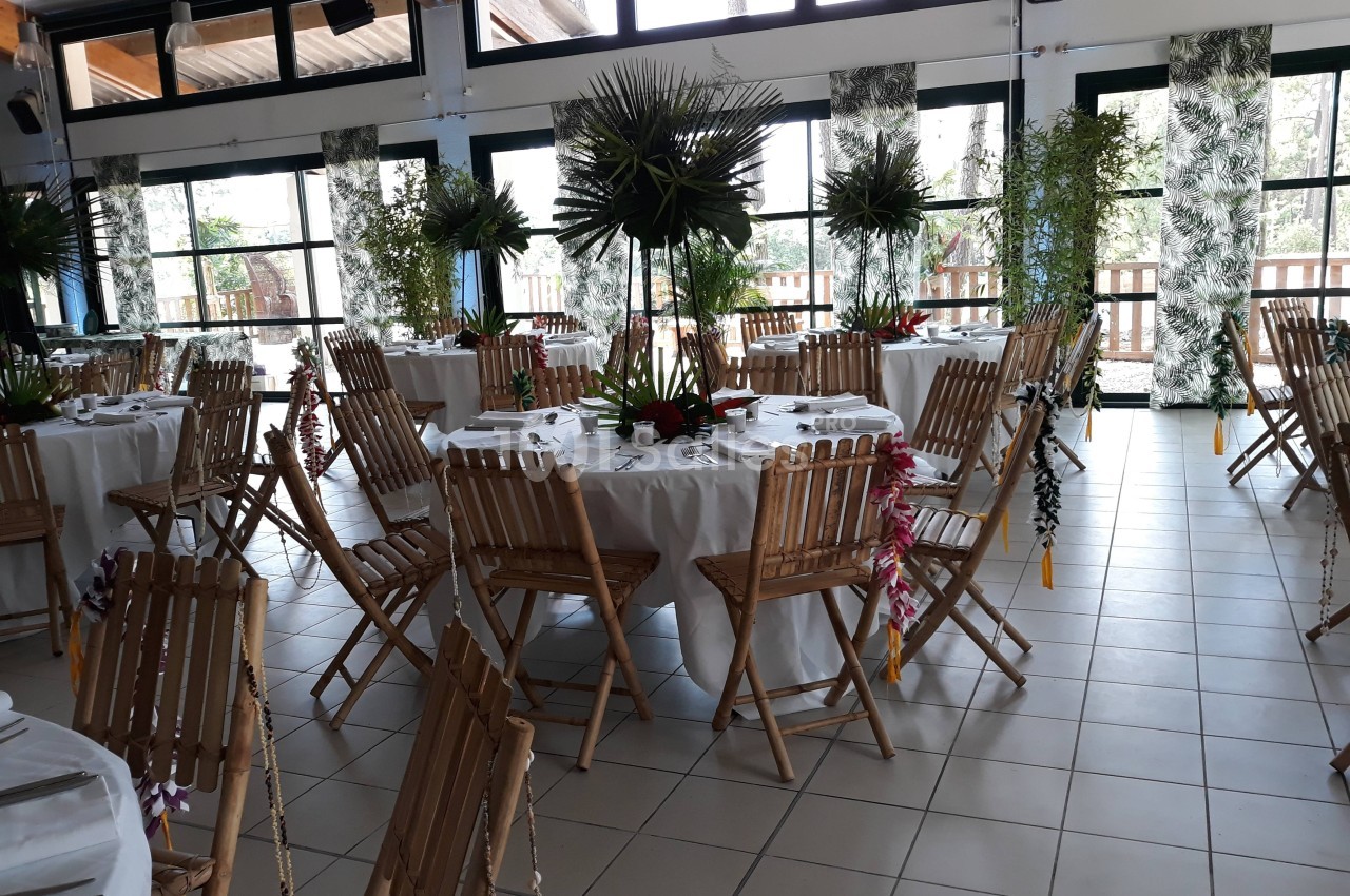 Salle décorée pour un événement, avec des tables rondes dressées, des chaises en bois et des arrangements floraux tropicaux.