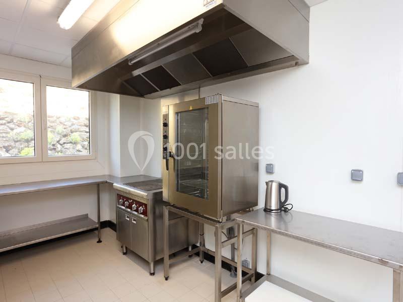 Cuisine professionnelle avec four en inox, plaque de cuisson, hotte, plan de travail en acier et bouilloire électrique.