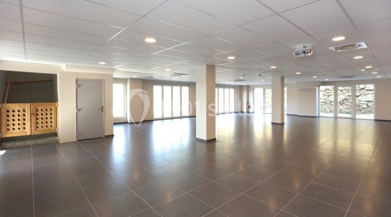 Salle spacieuse et lumineuse avec carrelage gris, murs clairs, colonnes et grandes baies vitrées.