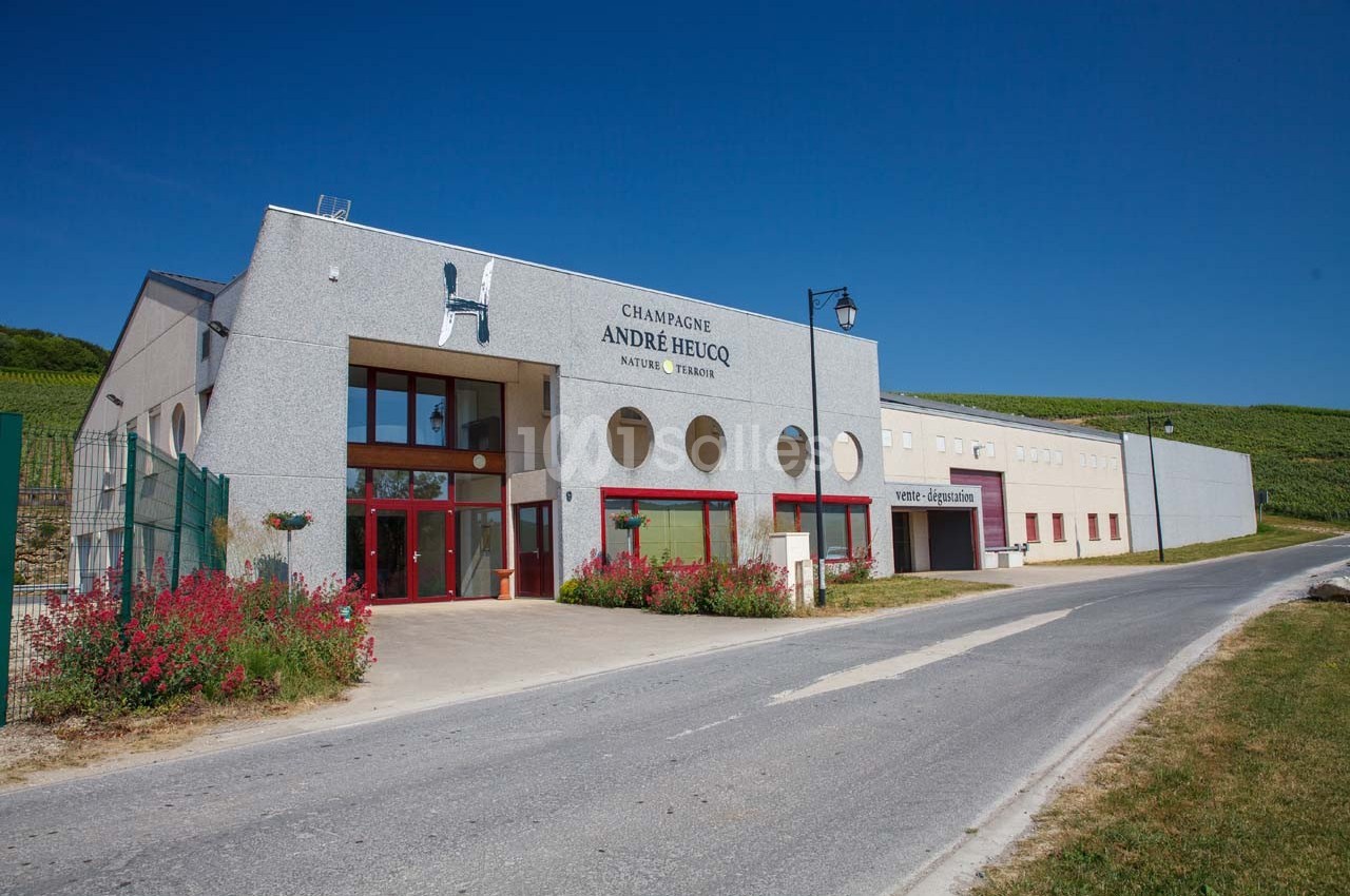 Bâtiment moderne du domaine viticole André Heucq, entouré de vignes, avec une route adjacente.