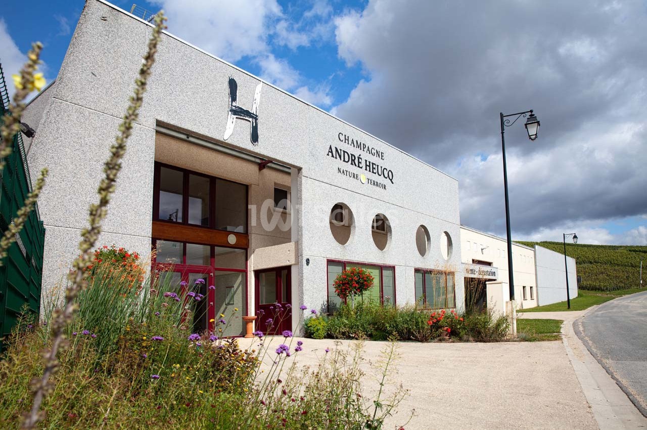 Façade d'un bâtiment viticole avec l'inscription ’Champagne André Heucq’, entouré de fleurs et de vignes.