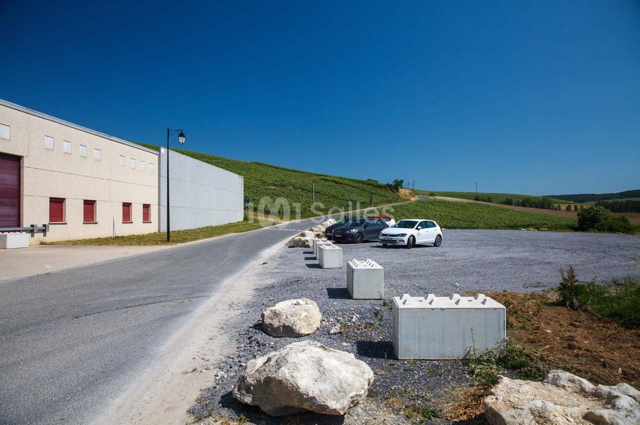 Parking avec quelques voitures stationnées près d'un bâtiment industriel, entouré de champs et d'un ciel dégagé.