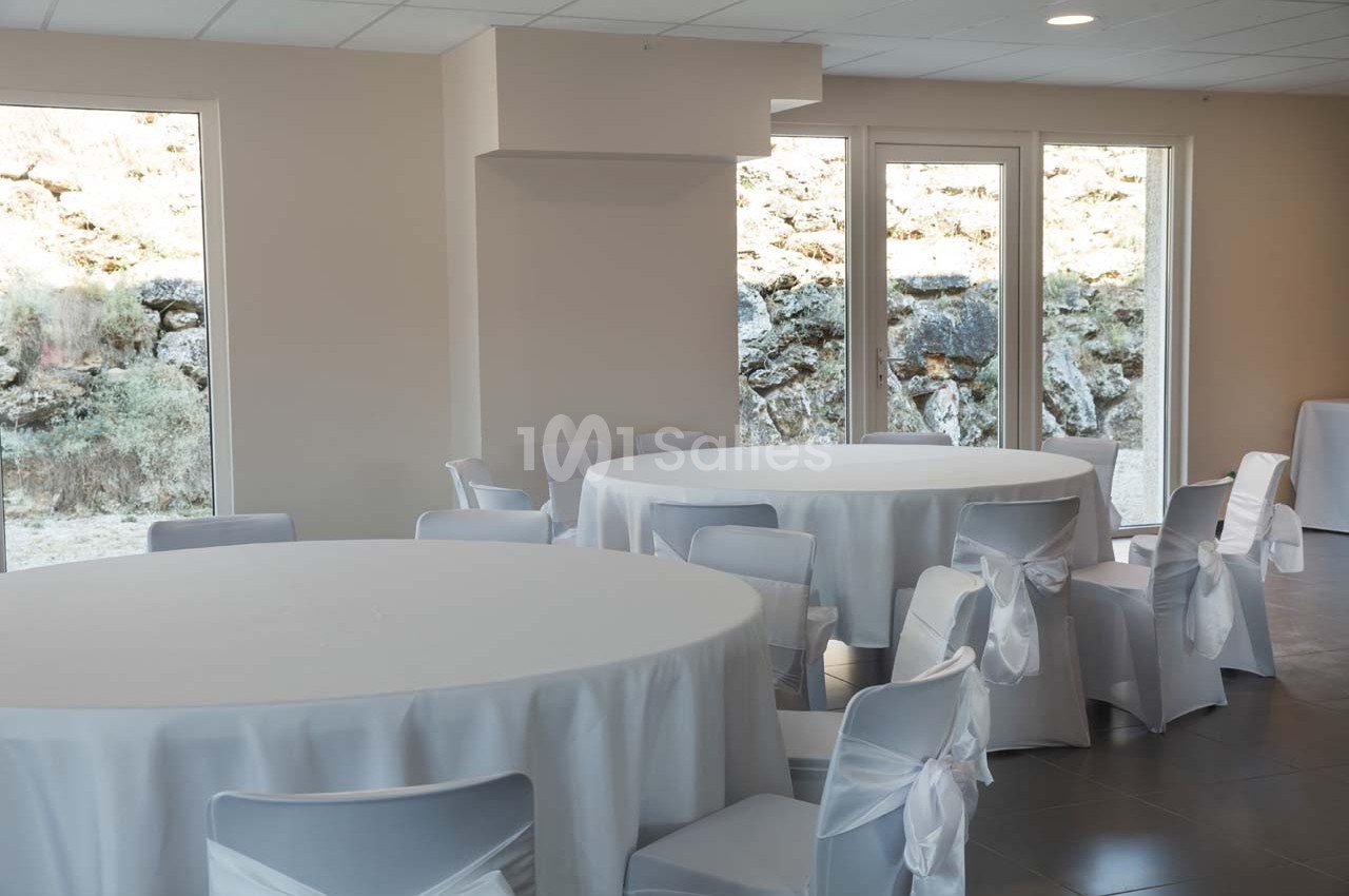 Salle lumineuse avec des tables rondes recouvertes de nappes blanches et des chaises décorées de nœuds.