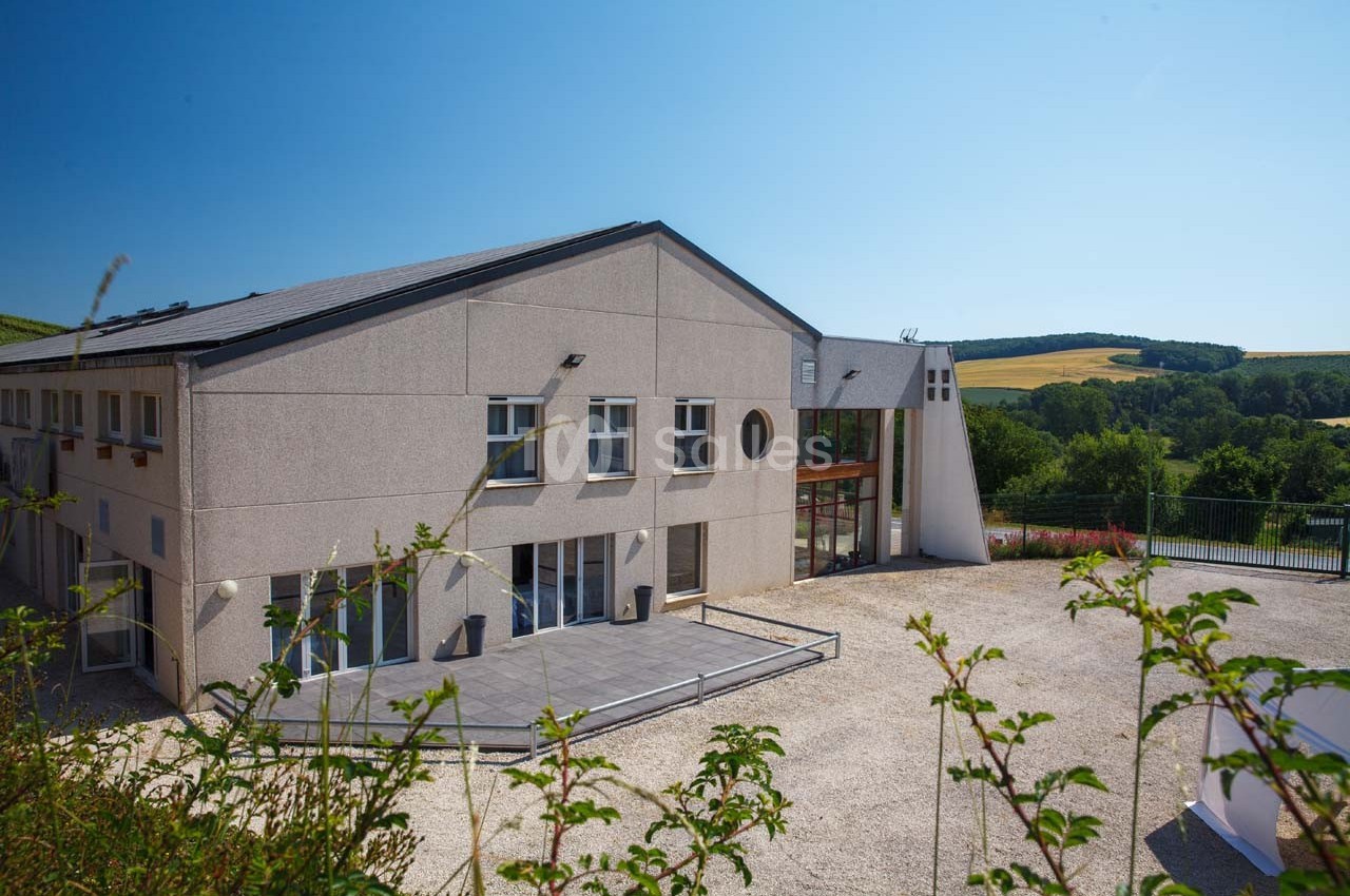 Bâtiment moderne avec façade beige, grandes baies vitrées et terrasse, entouré de végétation et d'un paysage rural.