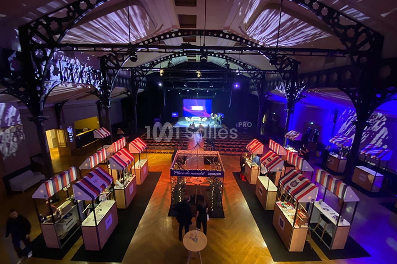 Vue d'une salle événementielle avec des stands colorés, un éclairage tamisé et une scène au fond.