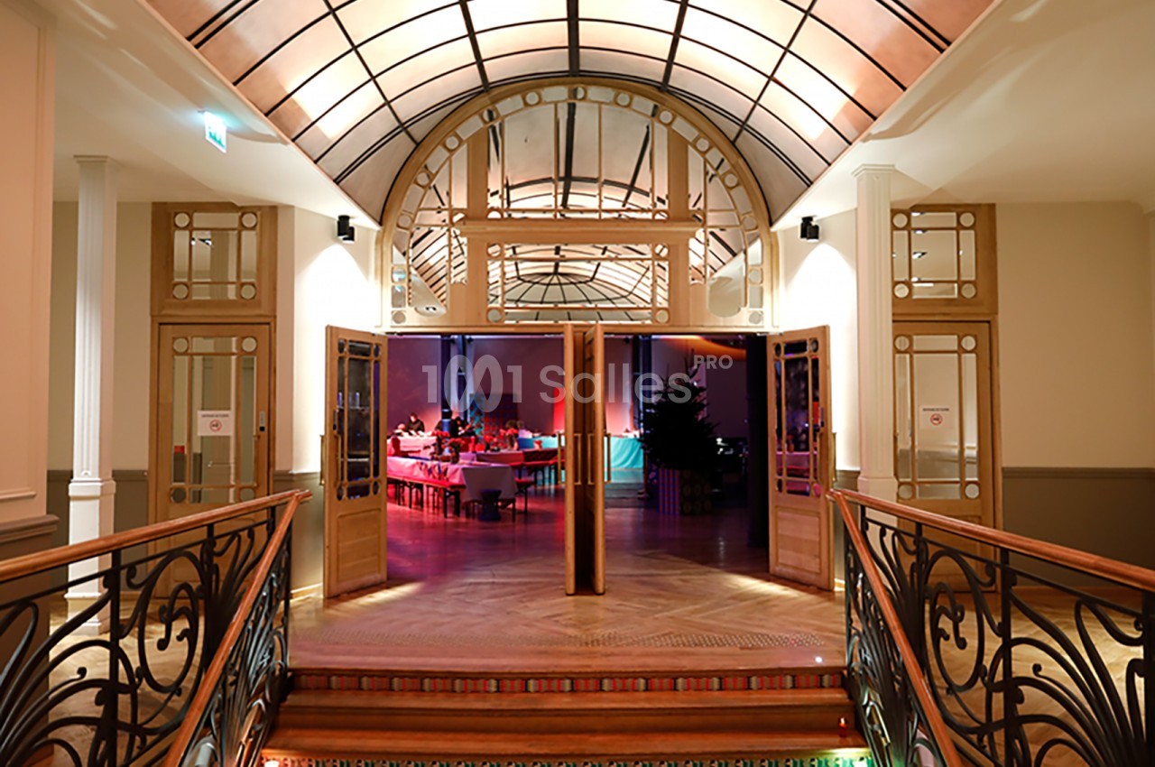 Vue d'une salle de réception éclairée, visible depuis un hall avec des portes ouvertes et un plafond voûté en verre.