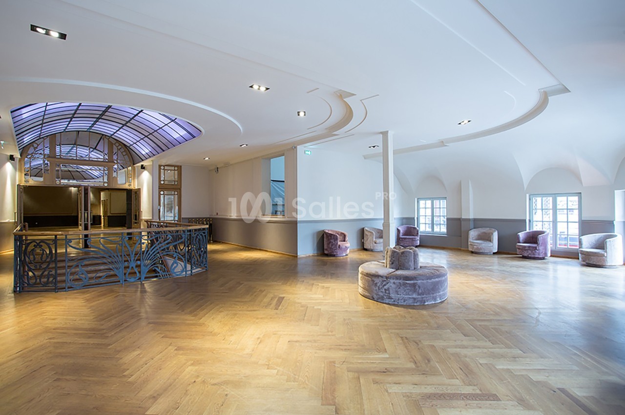 Salle lumineuse avec parquet en bois, fauteuils violets, balustrade décorative et plafond voûté avec verrière.