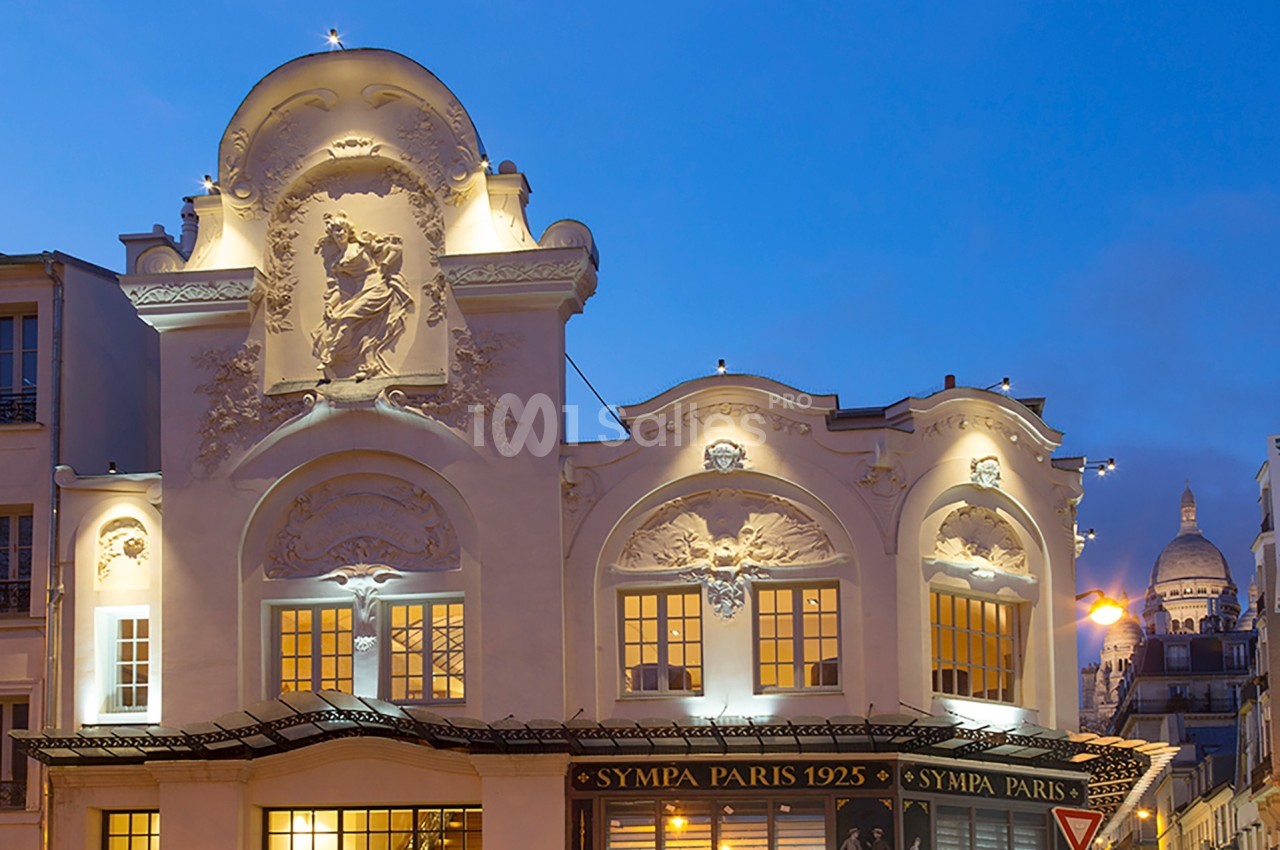 Façade d'un bâtiment parisien illuminé au crépuscule, avec des détails sculptés et le Sacré-Cœur visible en arrière-plan.