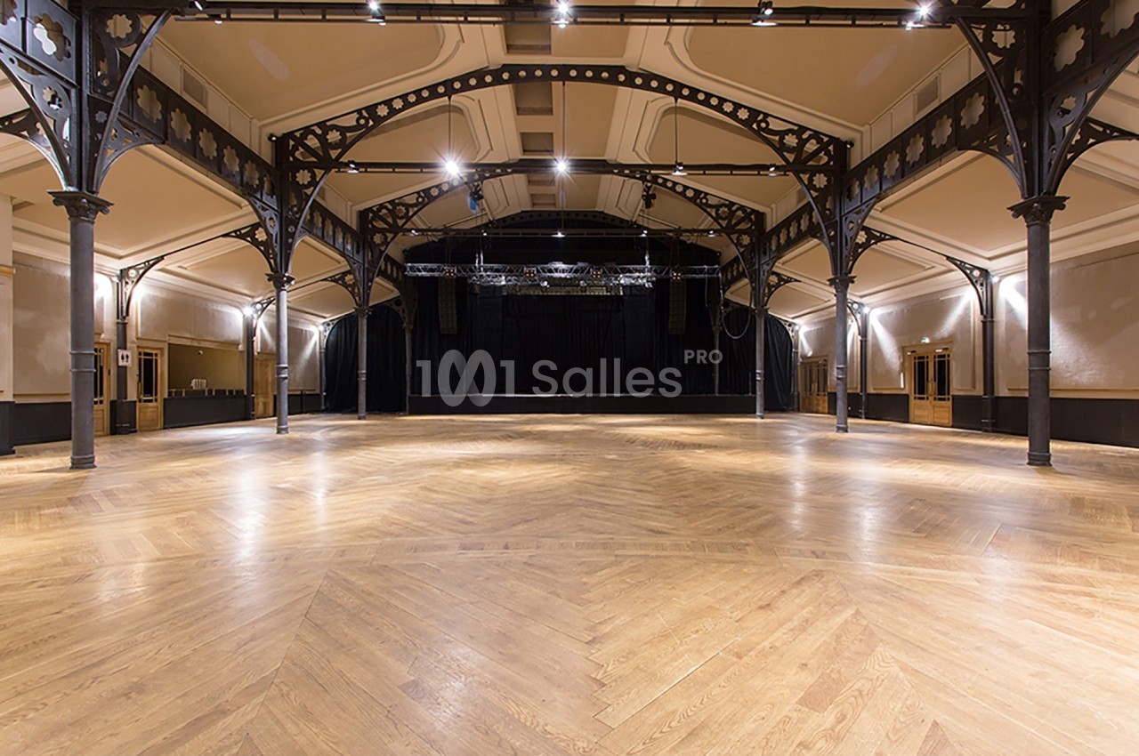 Salle spacieuse avec parquet en bois, poutres métalliques apparentes et scène noire en fond.