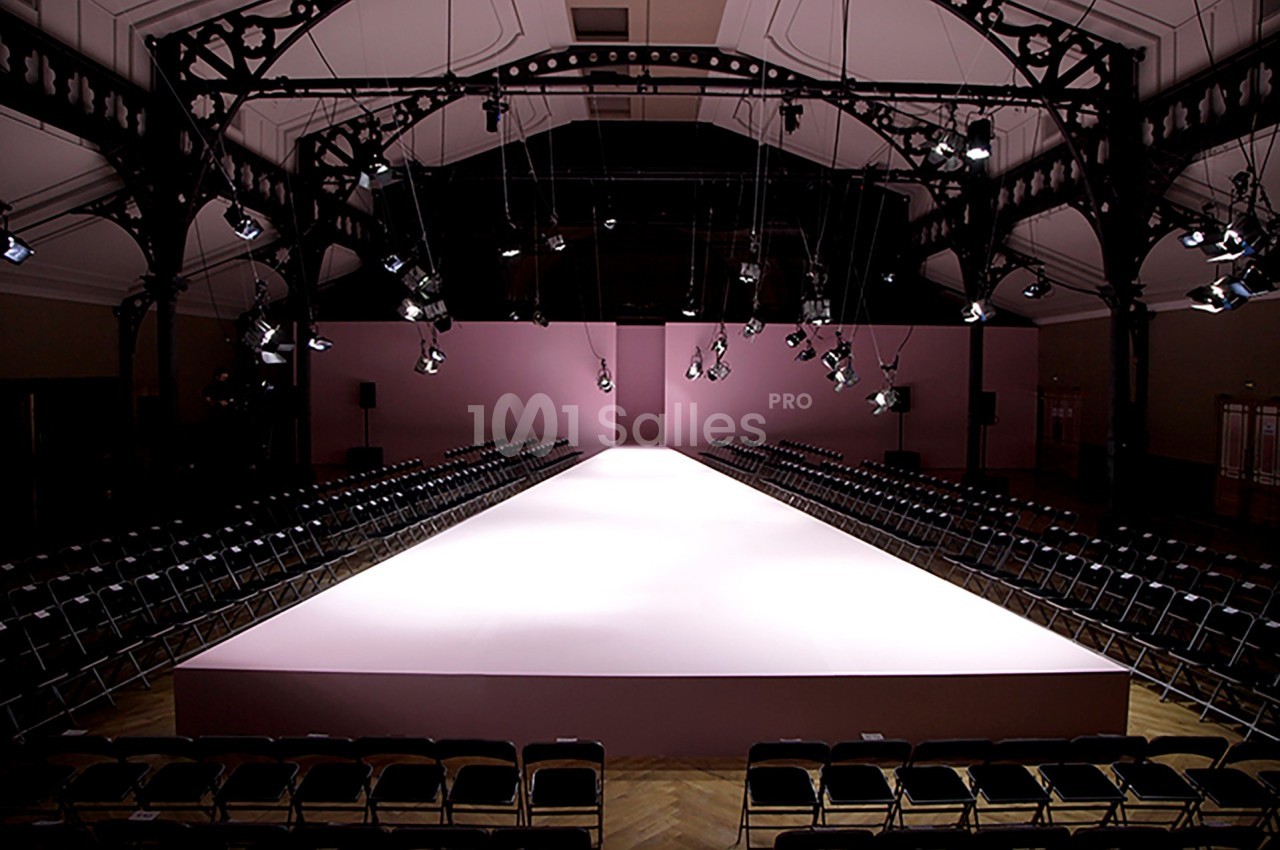 Podium de défilé de mode vide, éclairé par des projecteurs, entouré de rangées de chaises noires dans une grande salle.