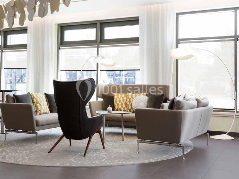 Salon moderne avec fauteuil noir, canapés gris, coussins décoratifs et lampadaires, éclairé par de grandes fenêtres.