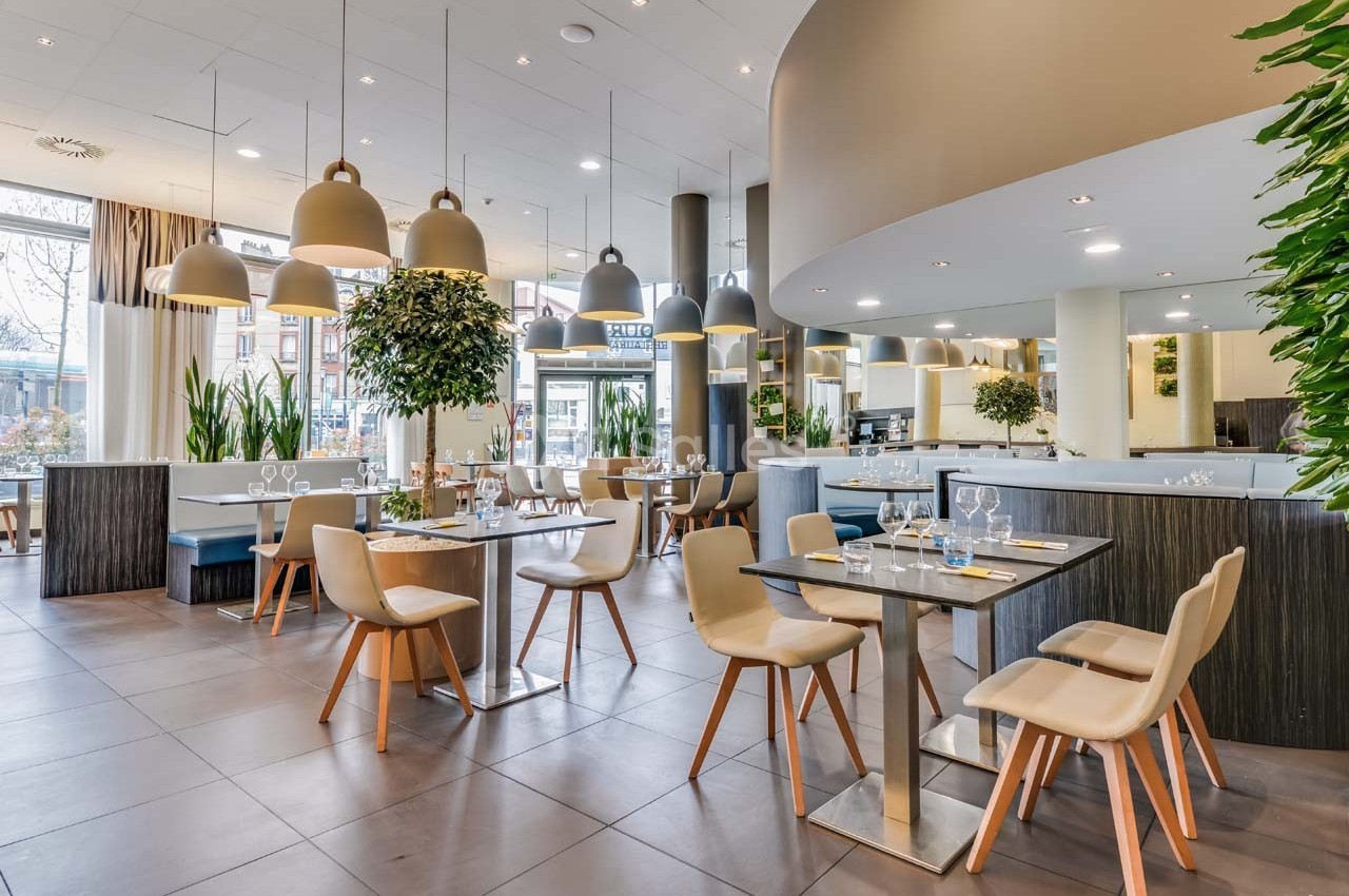 Salle de restaurant moderne avec tables dressées, chaises en bois clair, luminaires suspendus et plantes décoratives.